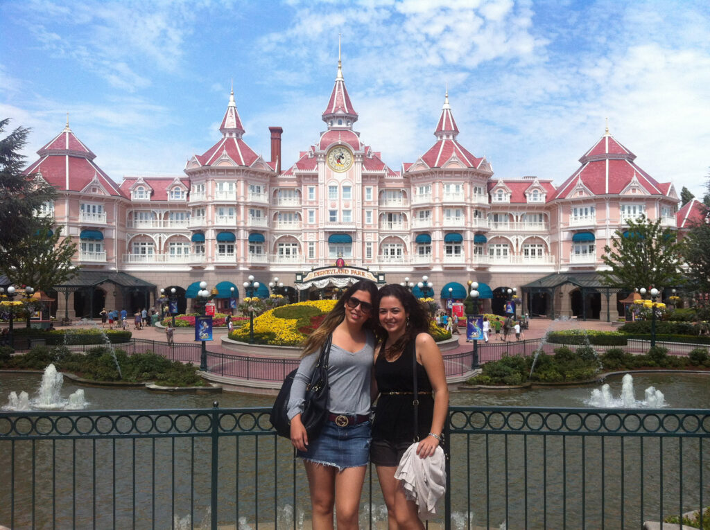 Entrada da Disney Paris, duas meninas em primeiro plano posam em frente a um castelo grande e cor de rosa que ocupa a foto de fora a fora e tem um lago na frente. 