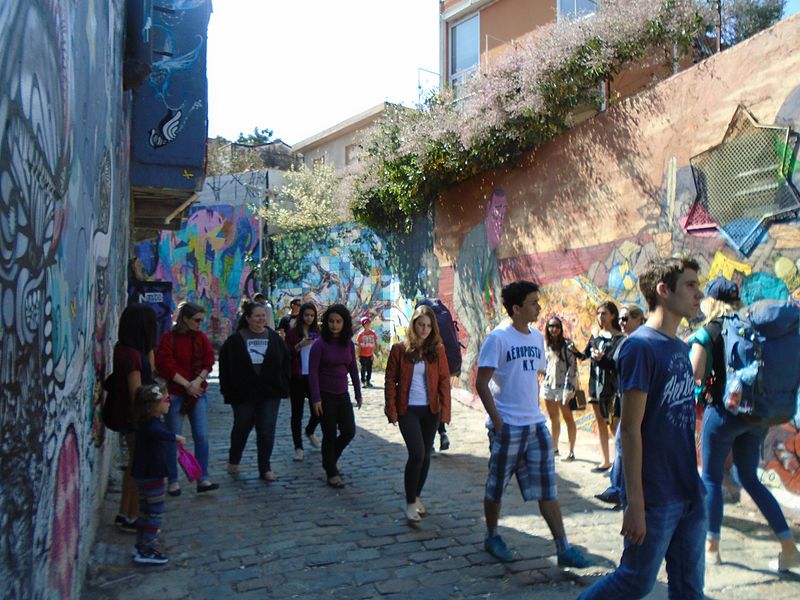 O que fazer em São Paulo? Ir ao Beco do Batman, na foto, uma ruela com dois muros,um em cada lado. Toda a extensão do muro está pintada com grafites e jovens caminham pelo pavimento de pedra.