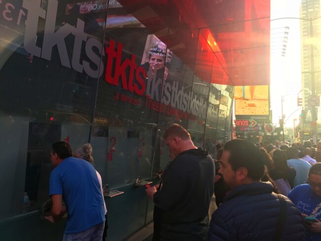 Foto de uma cabine de vidro onde se lê TKTS escrito em vermelho, e onde duas pessoas diferentes estão lado a lado comprando ingressos. 