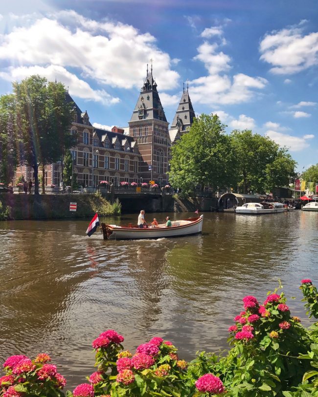 Barco cruzando um canal em Amsterdam, com flores e o museu Rijksmuseum ao fundo