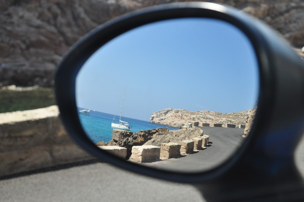 Foto de um espelho de carro. Refletido nele, um mar com um barco e uma estrada sinuosa.