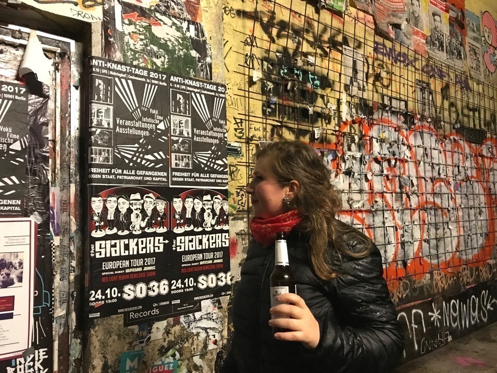 Mulher com uma garrafa de cerveja na mão, em frente a uma parede com várias figuras coladas em estilo lambe-lambe.