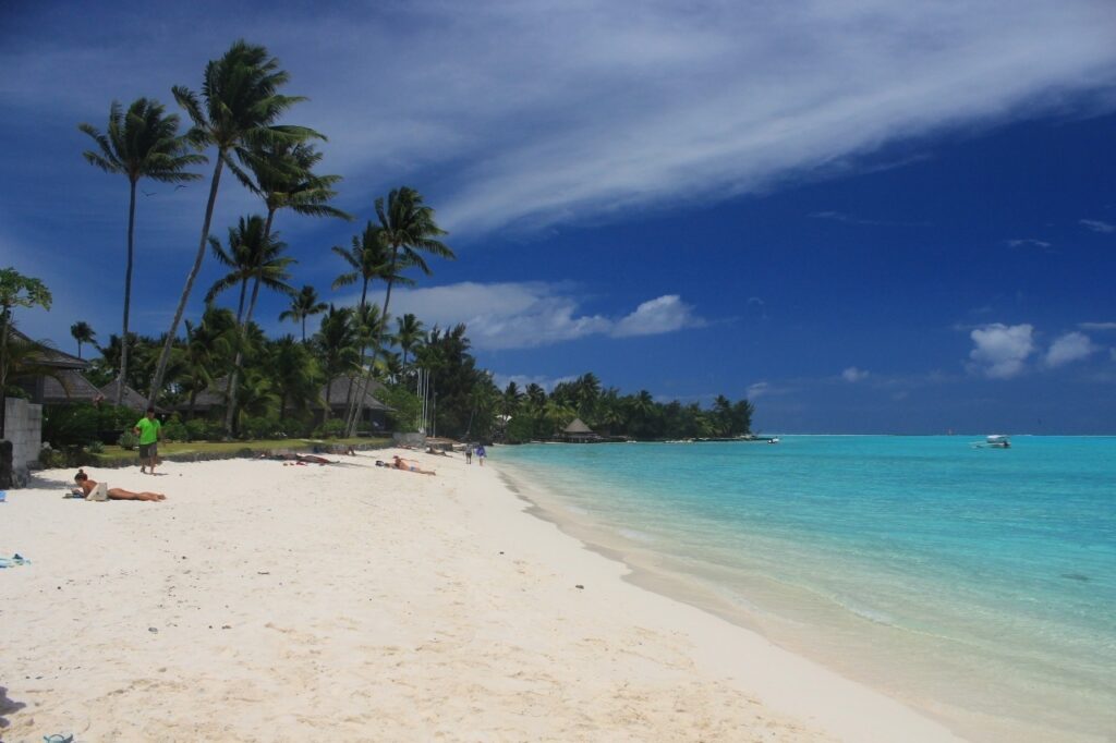 Praia em Bora-Bora, na Polinésia Francesa. Coqueiros cortam um céu azul, enquanto vemos abaixo uma areia branca e um mar azul turquesa