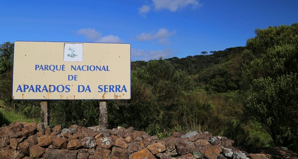 Placa bege, ligeiramente velha, onde se lê "Parque Nacional de Aparados da Serra" em Azul. A placa está sobre um muro de pedras e ao fundo há árvores bem verdes e um céu azul. 