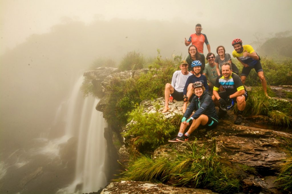 Clima ligeiramente neblinoso. A esquerda, uma cachoeira alta, e a direita, um grupo de pessoas sorri para a foto, sentados na mapa na borda da cachoeira ou em pé. 