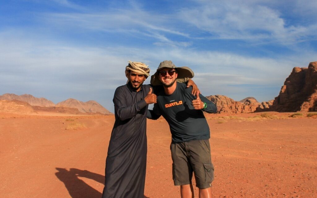 Guia Ahmed vestido com túnica cinza e turbante bege na cabeça, abraça homem com bermuda e camiseta e chapéu. O cenário de fundo é o deserto e as areias marrons.