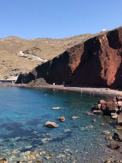 Foto de uma praia vista de frente que tem uma encosta avermelhada, areia com cor terrosa e um mar com diferentes tons de azul. É possível ver as pedras no fundo do mar e na direita da foto 