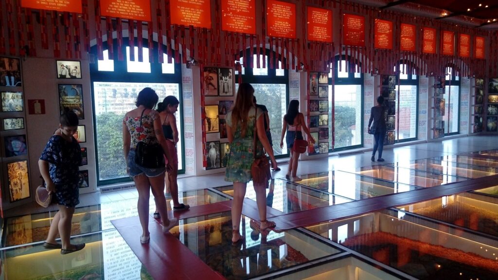 Pessoas caminham dentro de museu que tem 3 janelas grandes com uma decoração vermelha em cima.