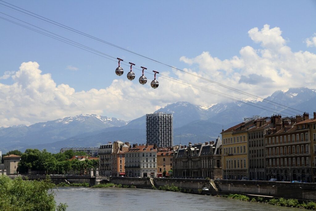 Vista do teleférico e do centro histórico da cidade de cima da Bastille em Grenoble. Montanhas ao fundo, um rio que cruza a cidade do lado esquerdo. O céu está azul e há 4 cabines sendo puxadas pelo teleférico.