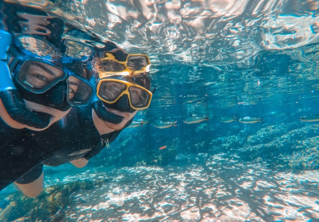 Casal submerso em uma água azul cristalina. Eles estão do lado esquerdo da foto com roupas de neoprene, máscara e snorkel e na metada direita da foto é possível ver algumas piraputangas e outros peixes.