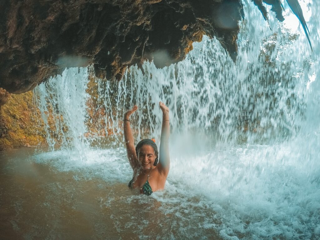 Foto tirada da parte de dentro da queda de uma cachoeira. Mulher com os braços para cima está bem abaixo de uma pequena queda d'água (menos de 2 metros de altura de queda).