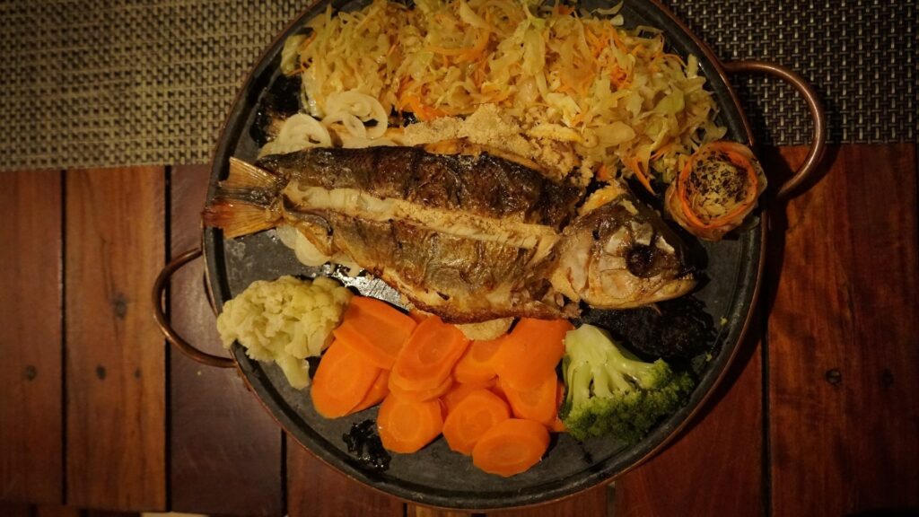 Foto tirada de cima. Grelha redonda comum peixe no centro, cenoura e legumes embaixo e saladinha na parte de cima.