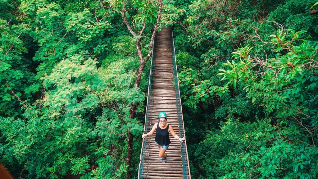 Mulher posa em cima de uma ponte de madeira no meio de uma selva com árvores bem verdes dos dois lados.