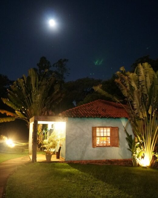 Casinha azul com dois coqueiros ao lado. É noite e a lua cheia está iluminada em cima da casa. 