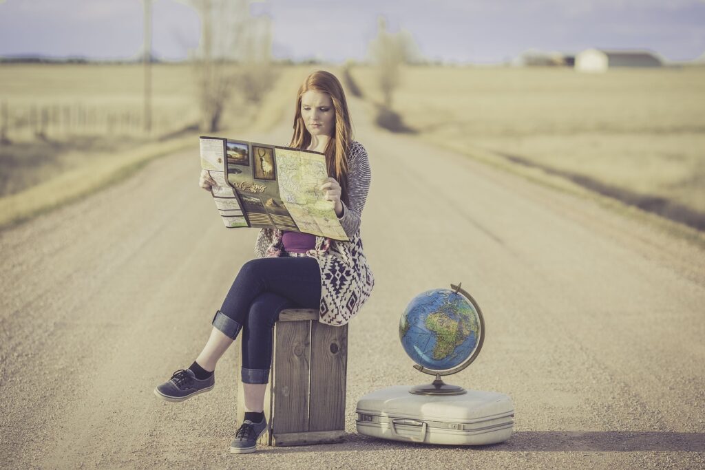 Bem no meio de uma estrada árida e vazia, uma mulher está sentada em cima da sua mala, com as pernas cruzadas olhando um mapa