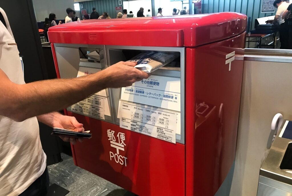 Uma viagem ao Japão precisa de internet. Na foto, uma caixa de correio de aproximadamente 50 centímetros de largura, onde um rapaz deposita um envelope pardo. Está escrito "Post" na caixa e mais textos em letras miúdas em japonês. 