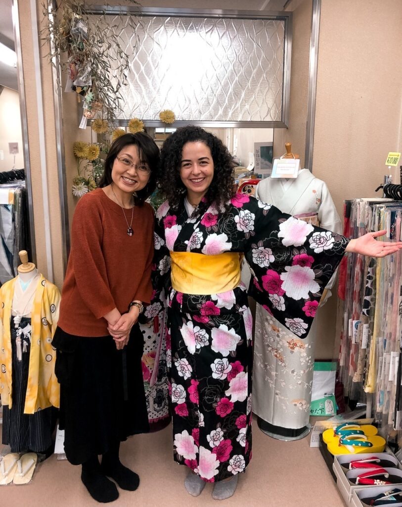 Em uma loja com tecidos e manequins vestindo kimonos ao fundo, há duas mulheres. A da esquerda está vestida com uma saia preta e blusa de manga comprida marrom avermelhada, e a da direita sou eu (Marcella) com os braços abertos e sorrindo vestindo um kimono de fundo preto com rosas grandes e cor de rosas, e uma faixa grossa amarela amarrada na cintura. 