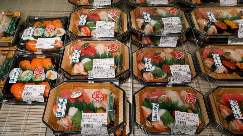 Foto com várias bandejas descartáveis dessas de comidas prontas. Dentro delas, sushis de peixes variados. 