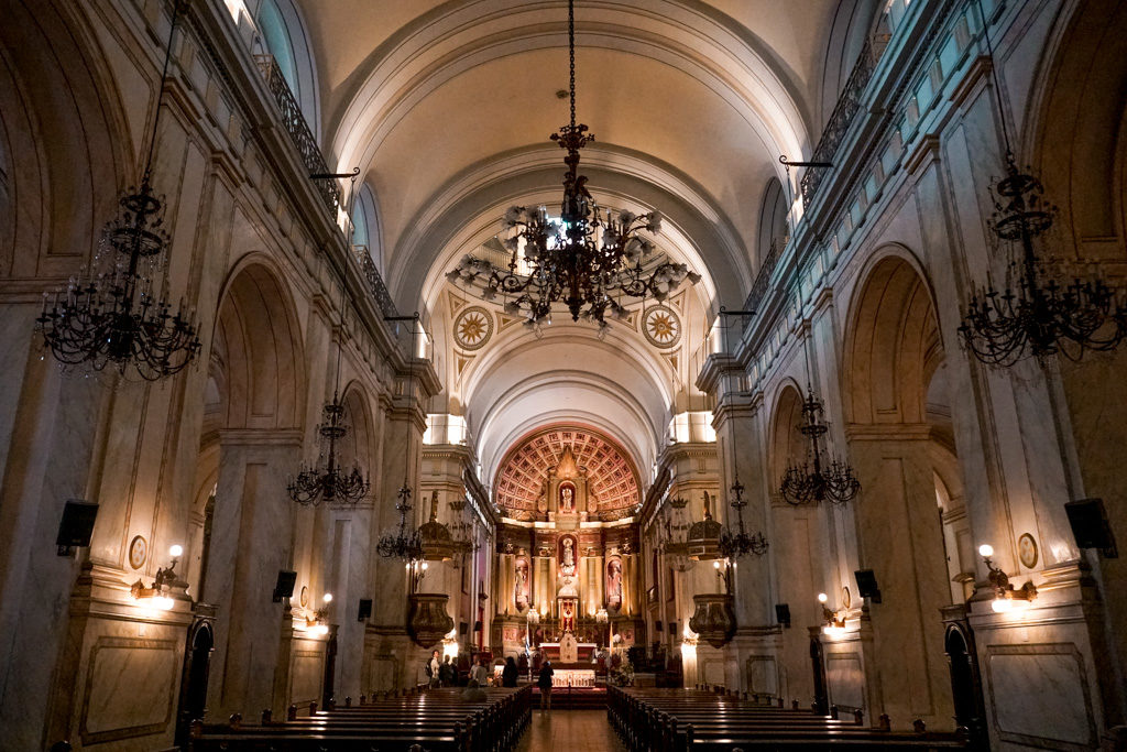 Interior clássico de uma igreja, com 3 lustres em cada lateral e um lustre maior no centro, um altar iluminado e com detalhes em madeira.