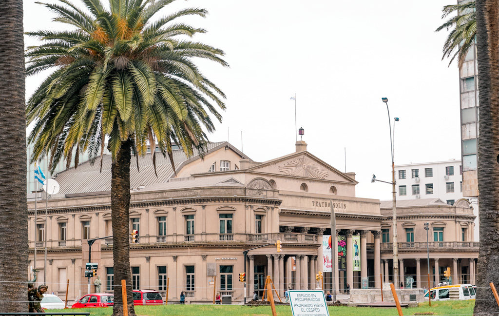 Foto da fachada clássica do Teatro Sólis em Montevidéu. Do lado esquerdo há um coqueiro e uma bandeira do Uruguai.