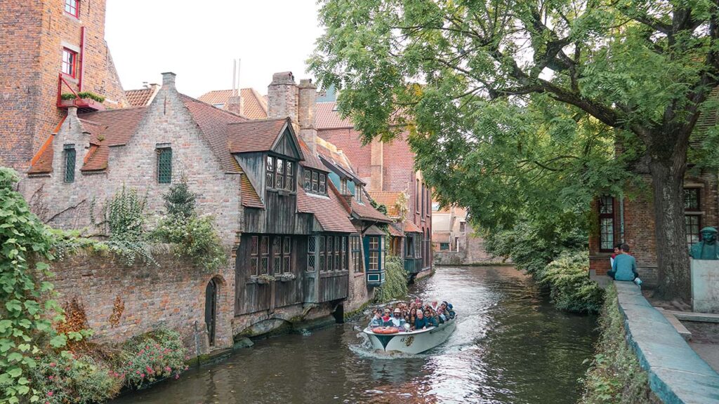 Barco atravessa um canal. A esquerda, na beira da água há casas medievais e a direita há uma árvore com folhas verdes.