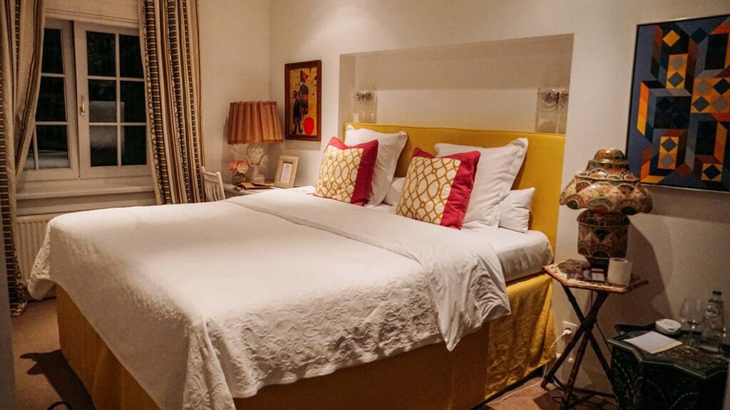 Foto de um quarto branco com uma janela a esquerda, com cortinas amarradas. Uma cama está no centro com lençois brancos e almofadas rodas e amarelo. 