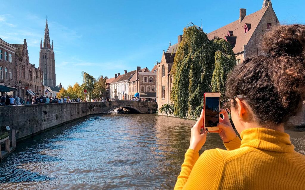 O cartão postal de Bruges. Mulher de camiseta gola alta amarela está de costas segurando um celular para tirar foto de um canal largo que parece um lago, com edifícios medievais ao fundo. Ela está no canto direito da foto. 