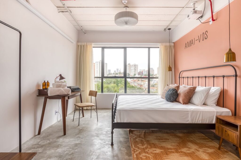 Imagem de quarto de hotel, com janelões ao fundo enquadrando uma cidade e verde das árvores, e uma parede rosa. A cama é de metal. Essa é a suíte do Selina Madalena, em São Paulo.