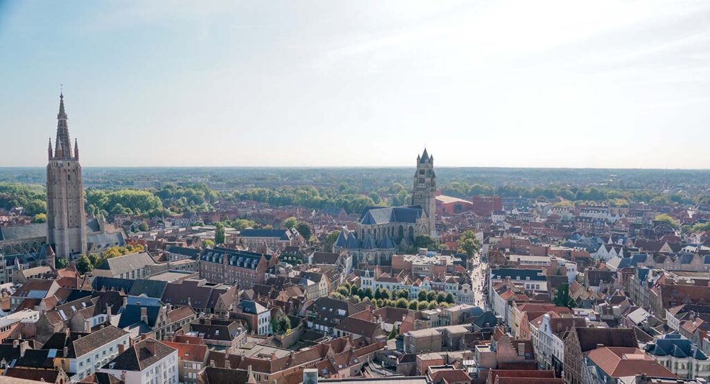 Vista panorâmica de Bruges, com suas torres de igrejas mais altas, e uma rua sinuosa com pedestres a direita. Bem ao fundo, há o verde dos campos. 