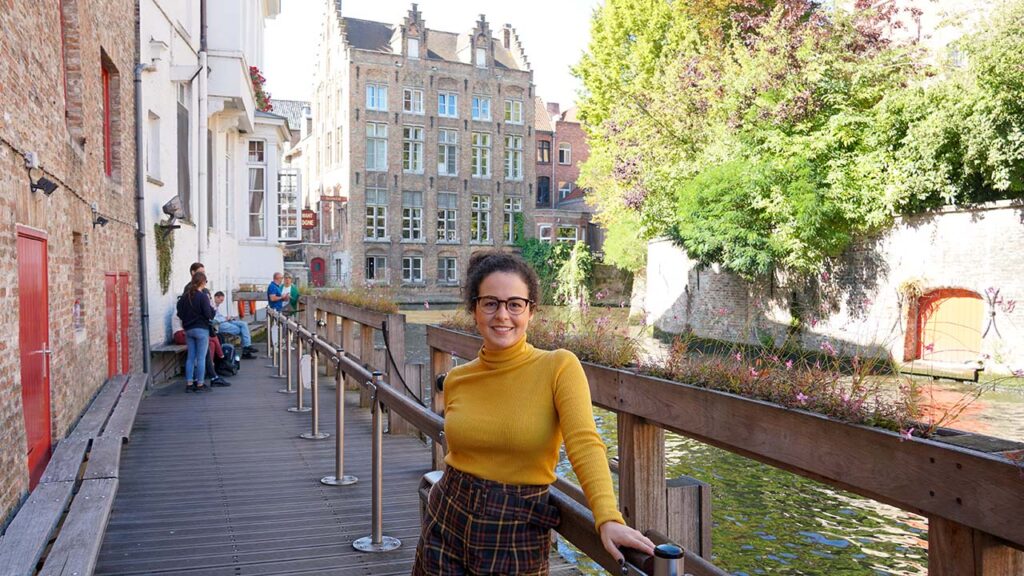 Mulher posa para foto com camiseta gola alta amerela e shorts marrom e amarelo xadrez. Ela está em um espaço de fila perto de um canal, de onde se pega o barco para o passeio pela cidade. 