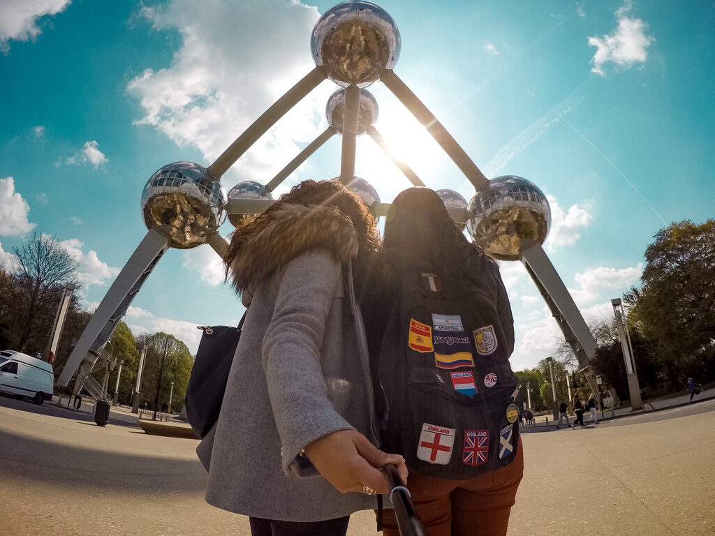 Foto de duas amigas de costas, uma com um casaco cinza e outra com uma mochila cheia de bandeiras de países costurada. Estão na frente de uma estrutura metálica em forma de átomo, em Bruxelas. 
