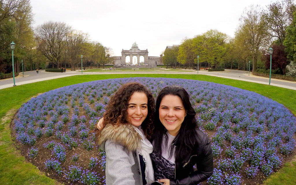 O que fazer em Bruxelas: Parque do Cinquentenário. duas amigas sorriem para a foto na frente de um canteiro grande de flores lilás. Ao fundo do parque há um edifício em forma de arco do triunfo. 