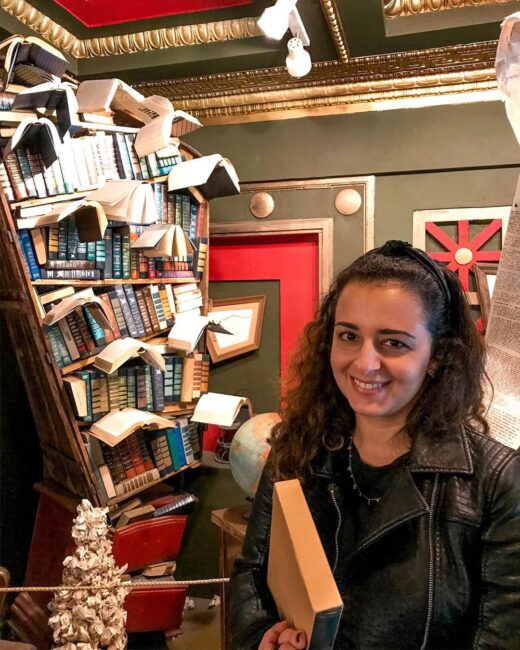 Mariana em livraria em frente a estante diferentona de livros e com um livro na mão
