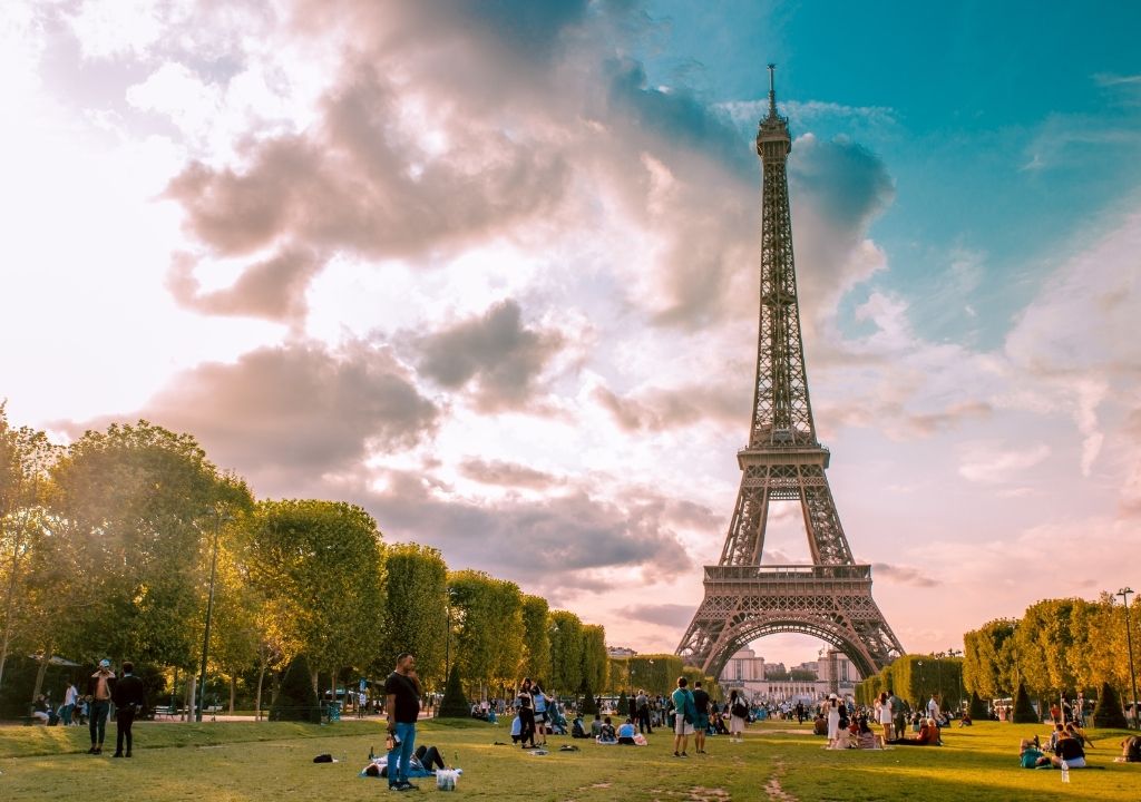 Torre Eiffel ao entardecer com céu rosado e várias pessoas fazendo piquenique no gramado diante dela