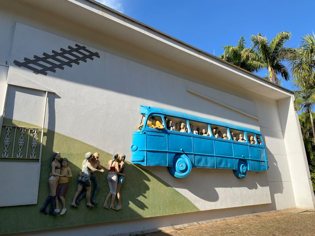 Inhotim: obra a céu aberto, a rodoviária de Brumadinho. Escultura na parede externa de uma galeria, com um onibus azul lotado de passageiros e 6 pessoas dançando forró do lado de fora. 