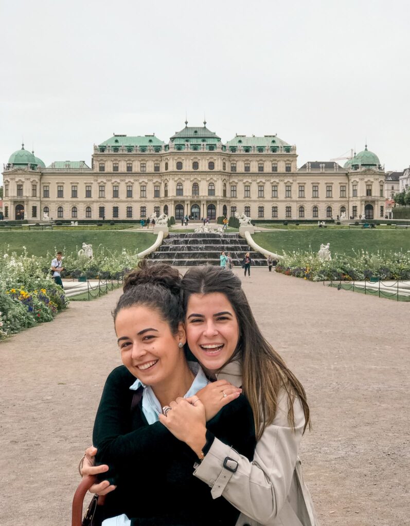 duas mulheres abraçam e sorriem para a foto na frente do palácio belvedere em viena