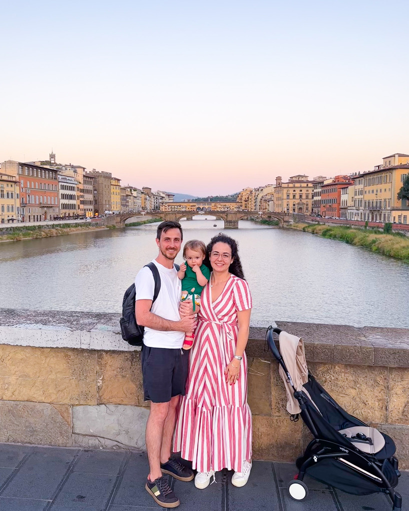 Casal segurando bebe posa em ponte em frente ao rio Arno, atrás está a Ponte Vecchio, em Florença. 